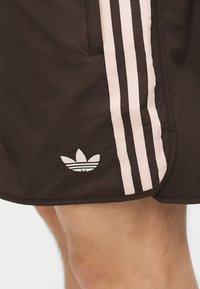 Κοντινή λήψη μαύρων σορτς Adidas με τρεις ροζ ρίγες και λογότυπο τριφύλλι στην πλευρά, που φοριούνται από άτομο με γυμνά πόδια.