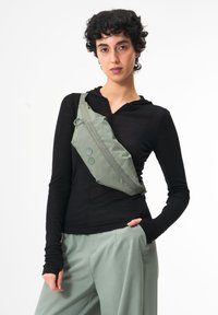 Grüner Umhängebeutel aus Stoff mit glatter Textur, mit einem geschwungenen Design und zwei runden Akzenten. Quer über den Körper getragen.