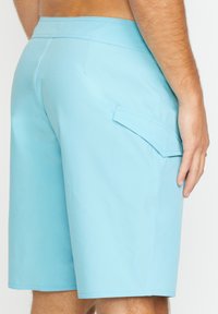 Shorts de bain bleu clair avec une texture lisse, une poche patch sur le côté et des coutures planes. La longueur atteint le mi-cuisse.