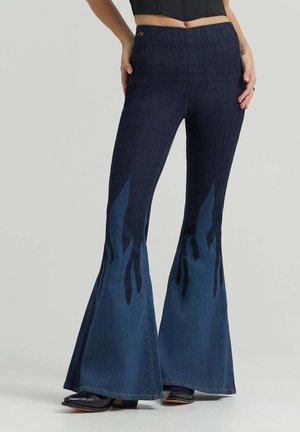 Frau trägt dunkelblaue Schlagjeans mit Flammenmustern an den unteren Beinen und schwarzen Stiefeletten, steht mit Händen in der Hüfte.