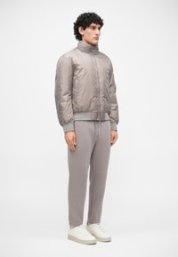Jachetă puf gri deschis cu guler înalt, țesătură texturată și închidere cu fermoar. Asortată cu pantaloni de trening gri și adidași albi.