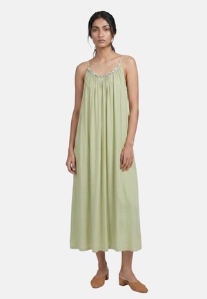 Femme portant une robe midi ample sans manches vert clair avec des bretelles spaghetti et des chaussures tissées marron à talons bas sur fond blanc.