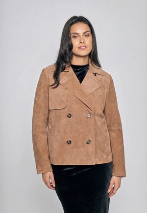 Kobieta w beżowej, dwurzędowej zamszowej kurtce na czarnej aksamitnej sukience, stojąca na tle jednolitego szarego tła.