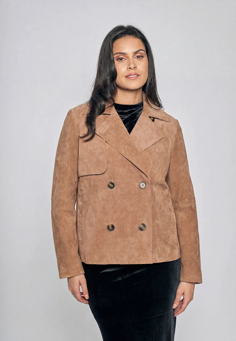 Donna che indossa una giacca doppiopetto in camoscio color sabbia su un abito in velluto nero, in piedi davanti a uno sfondo grigio uniforme.