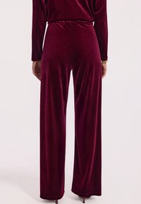 Kobieta w wysokich, szerokich spodniach z burgundowego aksamitu w talii, w komplecie z długim rękawem w tym samym kolorze, stojąca na jednolitym tle.