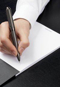 Czarny teksturowany długopis trzymany w ręce, piszący na białej, pustej kartce notatnika. Notatnik ma czarną skórzaną okładkę z widoczną marką.