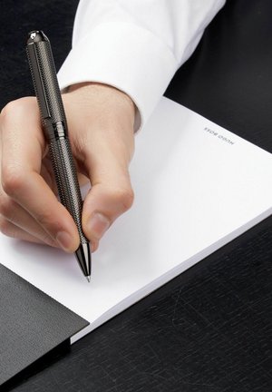 Črn teksturiran kemični svinčnik, ki ga drži roka, piše na praznem belem blokcu. Blokec ima črn usnjen ovitek z vidno blagovno znamko.