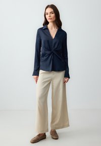 Navy Satinbluse mit geknoteter Vorderseite, langen Ärmeln und Kragen; kombiniert mit beigen weit geschnittenen Hosen und braunen Loafers.