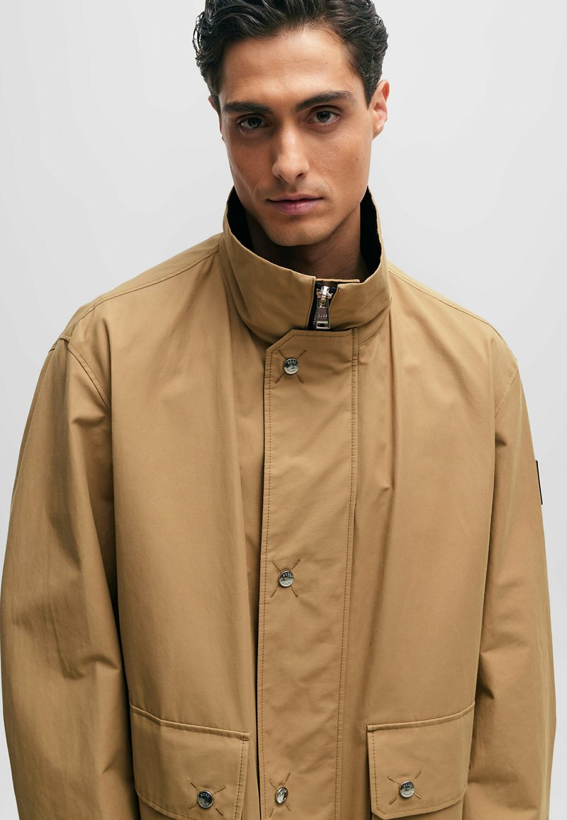 Beige Außenjacke mit hohem Kragen, Knopfverschluss, zwei vorderen Taschen und einer glatten Stoffoberfläche.