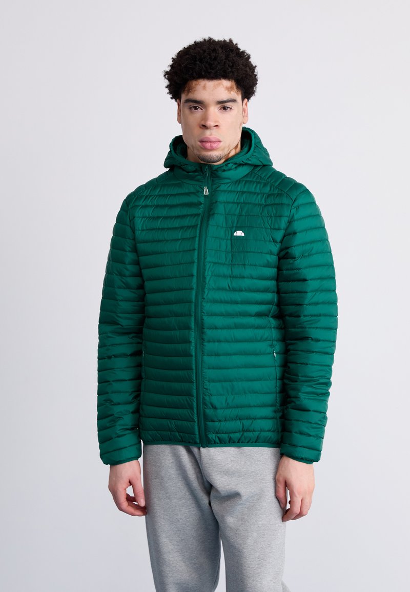 Chaqueta verde acolchada con capucha, con líneas acolchadas horizontales y un pequeño logo en el pecho. Combinada con pantalones de chándal grises.