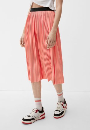 Person iført et koralfarvet plisseret midinederdel, hvid crop top, hvide sokker med pink striber og hvide sneakers med røde og sorte detaljer.