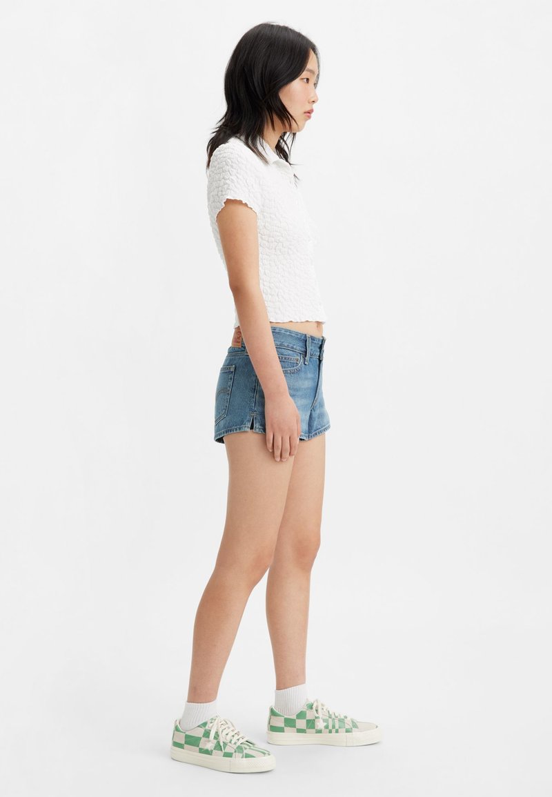 Haut court texturé blanc à manches courtes, associé à un short en denim bleu. Des baskets à carreaux blancs et verts complètent le look.