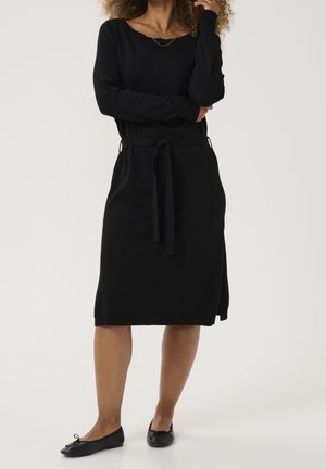 Femme portant une robe noire à manches longues, longueur genou, avec une ceinture à la taille et des ballerines noires, debout devant un fond uni.