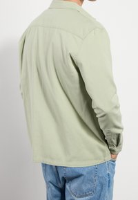 Persona vestida con una camiseta de manga larga verde claro y jeans azules de espaldas, con la mano derecha en el bolsillo, contra un fondo blanco liso.
