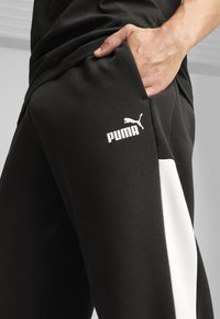 Schwarze Sporthose mit einem weißen Seitenpaneel, auf dem der Puma-Logo in Weiß auf dem Oberschenkel gedruckt ist und eine Hand in der Tasche steckt.