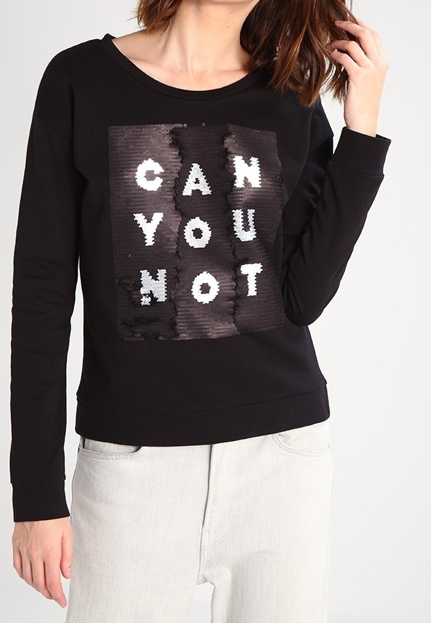 Sudadera negra con cuello redondo, que presenta un parche de lentejuelas texturizado con el texto "CAN YOU NOT" en colores contrastantes. Combinada con pantalones de color claro.