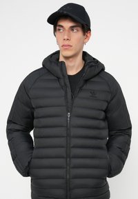 Melnā pūkā jacket ar kapuci, ar kvadrātu dizainu un rāvējslēdzēja priekšējo aizdari. Apdarināta ar melnu logo kreisajā krūtī un saskaņotu cepuri.