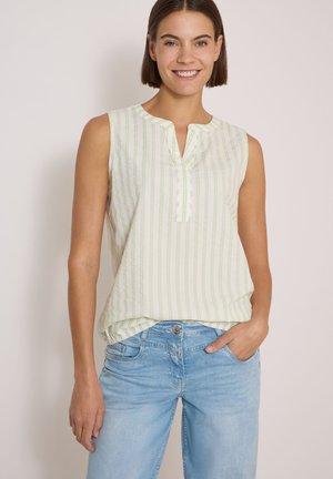 Jeune femme souriante, portant un haut blanc sans manches à rayures verticales vert clair et un jean bleu clair, debout devant un fond uni.