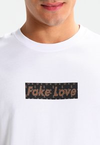 Hvit bomull t-skjorte med et rektangulært grafisk trykk i svart og brunt med teksten "Fake Love", omgitt av et mønstrete logodesign.