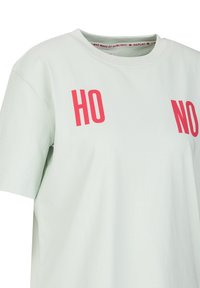 Lichtgroene katoenen t-shirt met ronde hals, korte mouwen en vetgedrukte rode tekst "HO" en "NO" op de voorkant. Vriendelijke textuur.