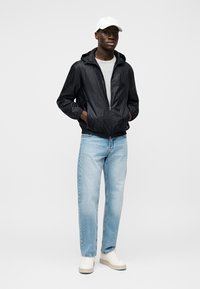 Geacă ușoară neagră cu fermoar, cu model texturat, tricou gri, blugi deschis la culoare și teniși albi. Modelul stă pe un fundal neutru.