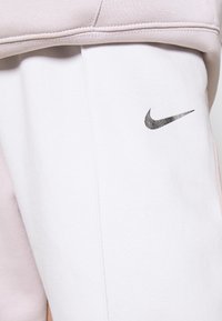 Nike sweatpants in white with a smooth finish, accented by a small black Swoosh logo on the thigh. Light pink sweatshirt partially visible above.