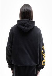 Sudadera negra con un ajuste holgado, presenta una gran capucha y texto amarillo en la manga. Fabricada con un tejido suave y texturizado para mayor comodidad.