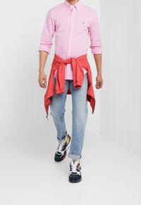 Světle růžová kostkovaná košile na knoflíky, červená bunda uvázaná kolem pasu, světlé modré džíny a vícerozměrné tenisky s vzorovaným designem.