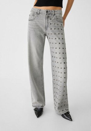 Kobieta nosząca jasnoszare szerokie jeansy z srebrnymi ćwiekami na prawej nogawce, w połączeniu z czarnymi butami na szpic i krótkim czarnym topem.