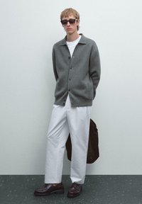 Grijze button-up cardigan, witte T-shirt, witte wijde broek, donkerbruine schoenen en zwarte zonnebril. Tas onder de arm gehouden.