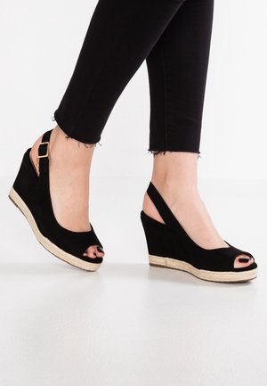 Pieds portant des sandales compensées noires à bout ouvert avec des brides à la cheville et des semelles plateforme beige tissées, associées à un pantalon noir raccourci.