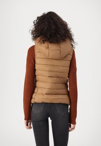 Gilet matelassé beige avec capuche amovible et manches orange côtelées. Présente des coutures horizontales et une coupe décontractée, associé à un jean foncé.
