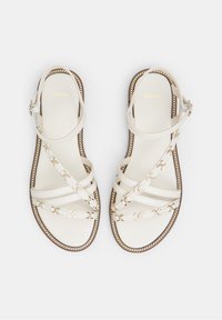 Valkoiset sandaalit, joissa on ristikkäiset hihnat synteettisestä materiaalista, kultasävytetyt ketjuakcentit ja litteä pohja, jossa on tummanvärinen reuna.