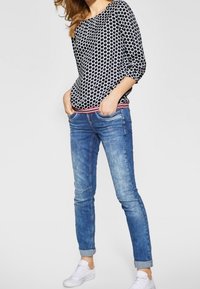 Kvinna klädd i en svart och vit prickig långärmad topp, blå skinny jeans med uppvikta byxben och vita sneakers, stående med händerna i fickorna.