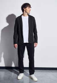 Svart kavaj med två fickor, bärs över en vit t-shirt. Tillsammans med svarta byxor och vita sneakers, stående mot en neutral bakgrund.