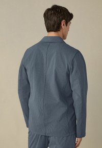Puuvillapuvust blazer tekstuuritellisessa sinisessä kankaassa, jossa on rento kaulus ja suora leikkaus, näkyvillä ompeluyksityiskohdilla ja sivutaskuilla.