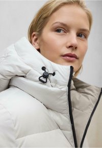 Chaqueta blanca acolchada con cuello alto, cremallera negra y toggle ajustable. Presenta una textura suave y lisa y líneas de diseño limpias.