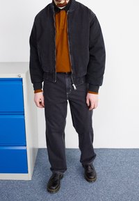 Czarna kurtka bomber z denimu, pomarańczowa koszulka polo, ciemnoszare jeansy i czarne sznurowane buty. Zauważalne detale w postaci zamka błyskawicznego i kontrastowego koloru na ramionach.