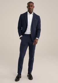 Costume à carreaux navy avec une veste sur mesure et un pantalon slim. Chemise habillée blanche en dessous, complétée par des chaussures noires à pampilles.