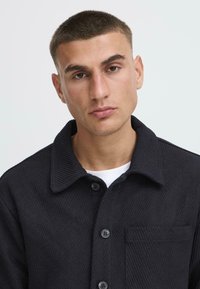 Black corduroy jacket with a collared neck, button closure, and a chest pocket. Textured fabric with vertical ribbing.