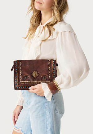 Vrouw in een witte blouse met ruches en licht gerafelde jeans houdt een bruine leren clutch vast met gouden en turquoise studs en een decoratieve centrale versiering.