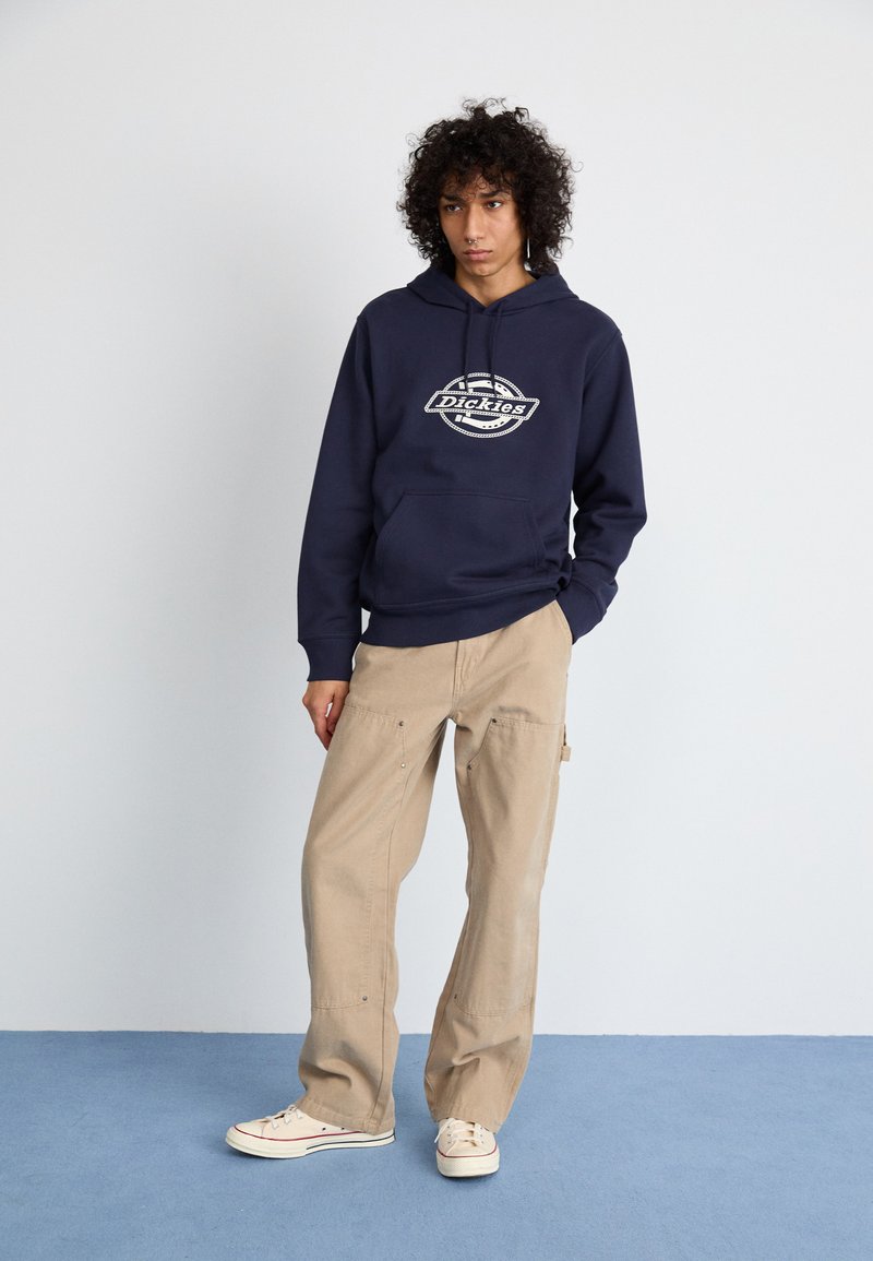 Felpa blu navy con logo, tasca a marsupio; pantaloni cargo beige con tasche laterali e cuciture sottili; sneakers crema con dettagli rossi.
