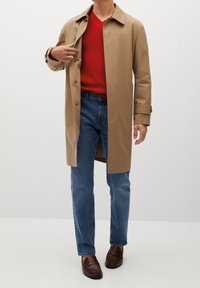 Helle beige Trenchcoat mit großen Knöpfen, rotem geripptem Pullover, blauen Jeans und braunen Loafers. Glatter Stoff und taillierte Passform.