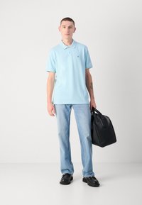 Lyseblå polo shirt, korte ærmer, sort logo på brystet; parret med lyseblå jeans og sort lædertaske; sorte sko, stående pose.