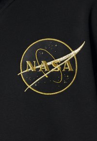 Černá tkanina má vyšívané logo NASA v zlaté barvě, s kruhovým designem, hvězdami a zakřivením, které představuje trajektorii vesmírné lodi.