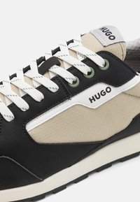 Baskets noires et beiges en tissu texturé, avec des lacets blancs, une semelle en caoutchouc et un logo "HUGO" sur le côté et la languette.