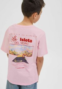 Gaiši rozā T-krekls ar grafisku druku, kurā ir "La Isleta Del Moro". Aizmugurē ir ainavas māksla un teksts. Mīksts kokvilnas materiāls.