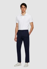 Polo shirt bianco a maniche corte con colletto, abbinato a pantaloni blu navy e sneakers bianche, presentato su uno sfondo grigio uniforme.