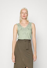 Top sem mangas de verde claro com bordado floral no decote em V, combinado com uma saia envolvente castanha com um detalhe de cintura amarrada.