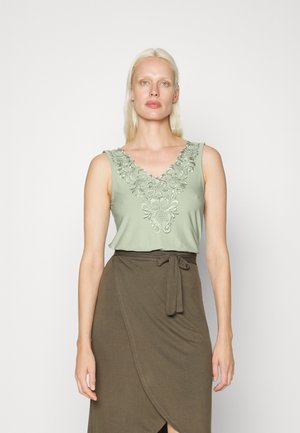 Top sem mangas de verde claro com bordado floral no decote em V, combinado com uma saia envolvente castanha com um detalhe de cintura amarrada.
